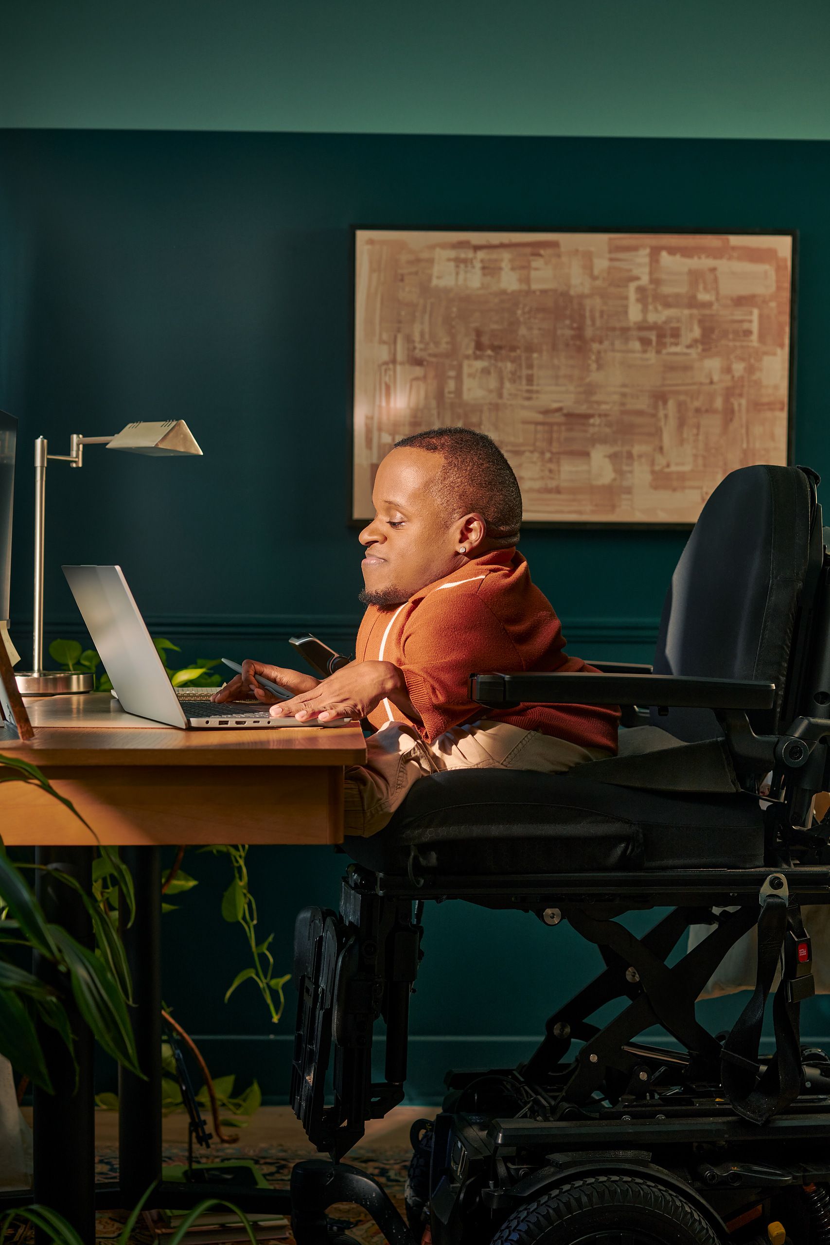 A black man who is a little person is seated in his power wheelchair in a dark teal home office. He types on a laptop on his desk. A lamp and a framed photo are on the desk in front of him. His wheelchair is in an elevated position, allowing him to be level with his desk. His desk is surrounded by several house plants and an abstract art piece. He has closely cropped black hair, brown eyes, and is wearing a rust-orange knit quarter-zip sweater with white vertical stripes, light khaki trousers, and a small stud earring. A tote bag hangs on the back of his wheelchair.