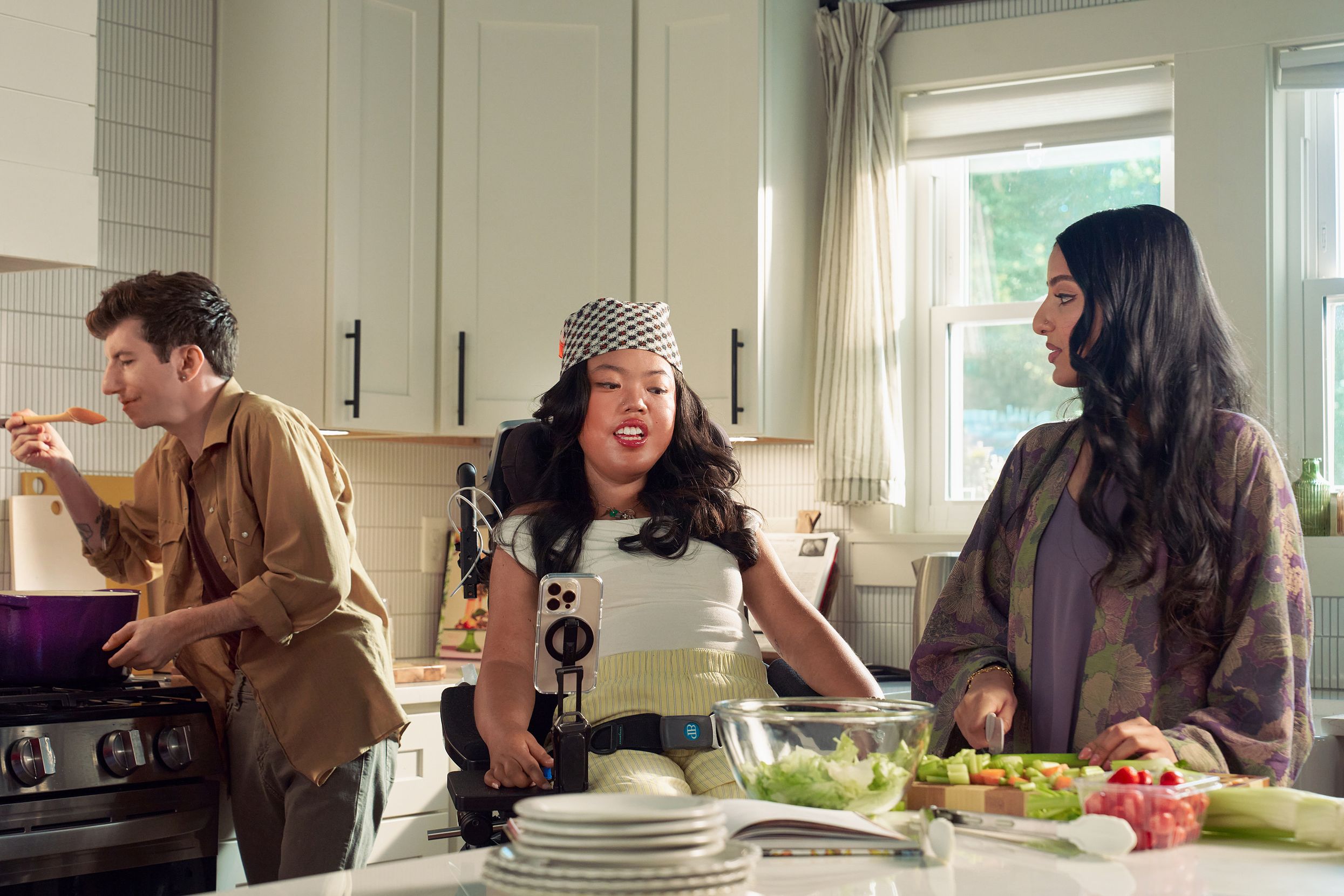 A SouthEast Asian woman with curled black hair seated in her power wheelchair gives her roommate instructions on how to prepare a salad in their bright kitchen. Her smart phone is mounted to her wheelchair. Behind them, a white male with Moebius Syndrome and a face that is paralyzed holds a wooden spoon up to his lips to taste a soup on the stove. The SouthEast Asian woman is wearing a white top with striped, yellow linen pants and a checkered headscarf. He has short, wavy, reddish brown hair and is wearing a tan button down shirt with the sleeves rolled up, showing arm tattoos. Their roommate has long dark hair and is wearing a purple floral dress.