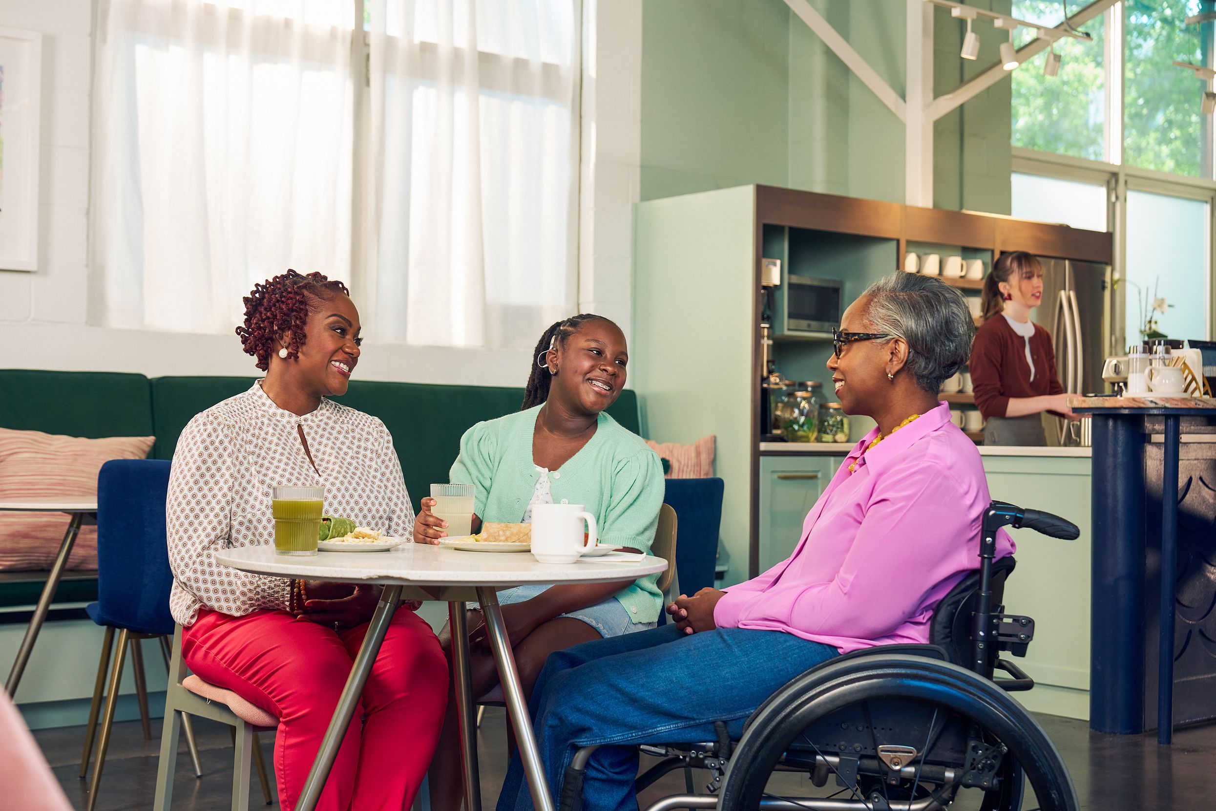 An African American mother, daughter, and auntie are smiling while chatting at a table in a modern cafe. The auntie is paraplegic and is seated in a manual wheelchair. She has short gray hair is wearing a lavender silk blouse and dark blue jeans. The mother has short tightly curled red hair and is wearing a patterned blouse and red pants. The daughter is wearing a cochlear implant and has her braided hair pulled back into a pony tail. She is wearing a light green cardigan and jean shorts. Behind them, a barista prepares drinks and a man types on a laptop.