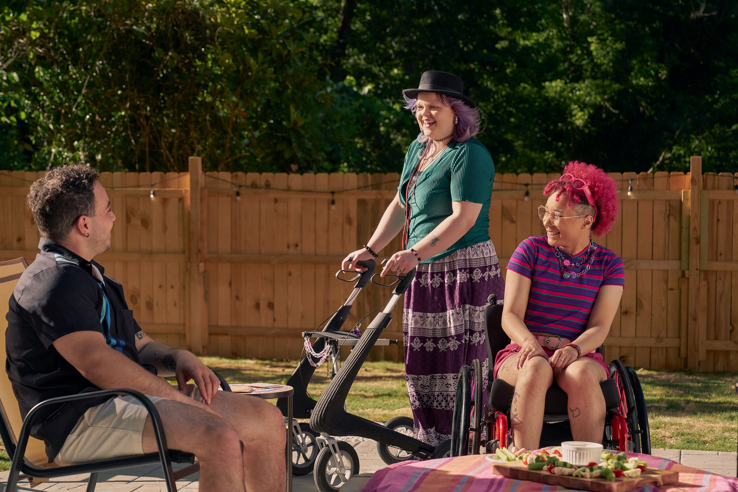 A white nonbinary person stands with their uniquely shaped dark gray rollator that is decorated with purple acrylic chains. They have purple layered hair with two long thin red braids on both sides and are wearing a black hat, green shirt, and purple patterned skirt. They chat with a friend, who is a multi-ethnic man with curly black hair, who is sitting in a patio chair. Next to them is a Black nonbinary person seated in their manual wheelchair with rainbow spokes. They have a curly pink fro with loc bangs and are wearing bright pink sunglassese on their head, a purple and pink striped crop top, pink shorts, and purple sandals. They have several tattoos on both legs.