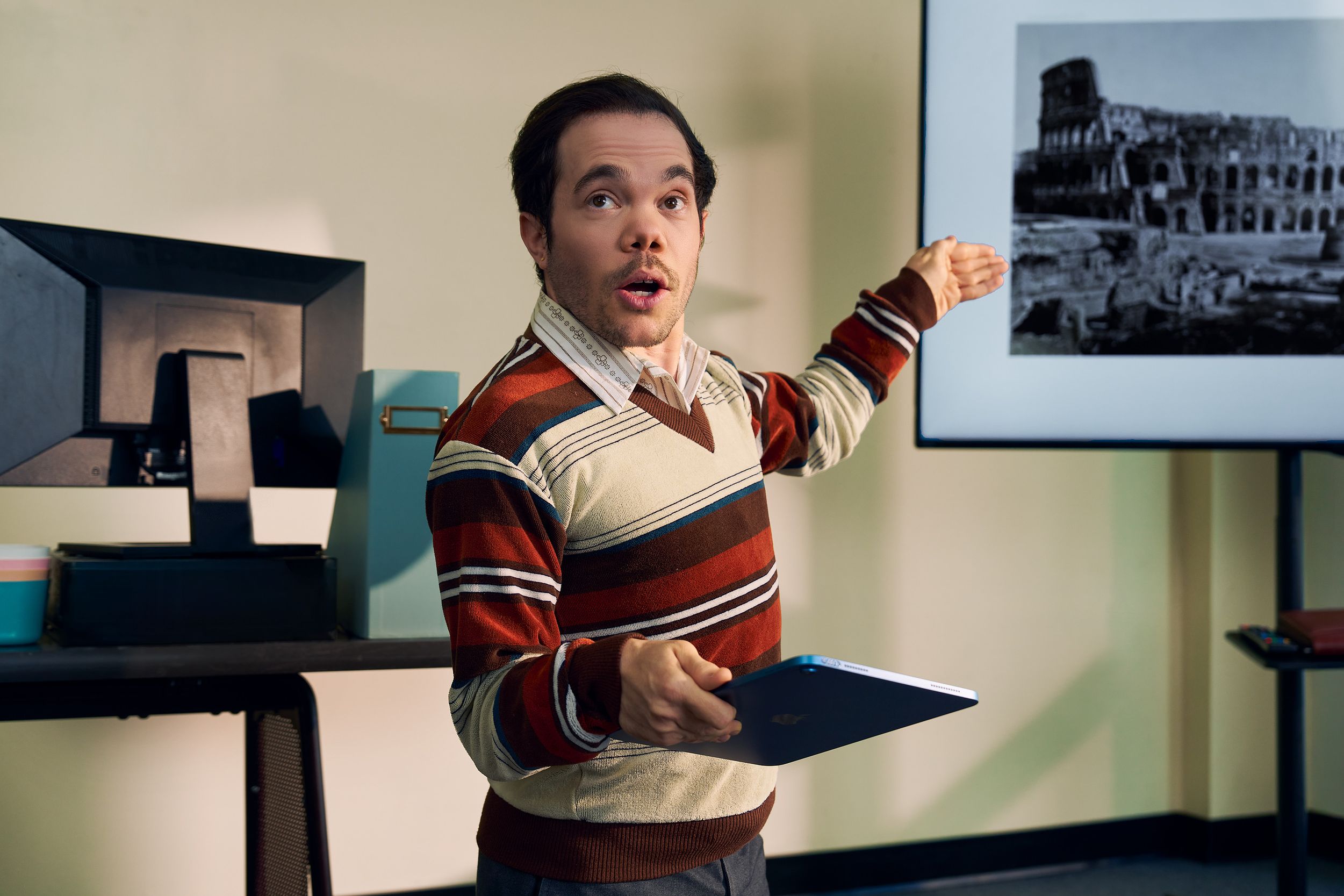 A white male professor with medium brown hair and light brown eyes who has dwarfism gestures at a large monitor next to him, displaying a black and white photo of the Colosseum. He is holding a tablet in his other hand. He is dressed in a striped V-neck sweater, charcoal dress pants, and dressy-casual shoes, standing with a warm, confident presence. Behind him, his desk has a computer, a file holder, and a mug.