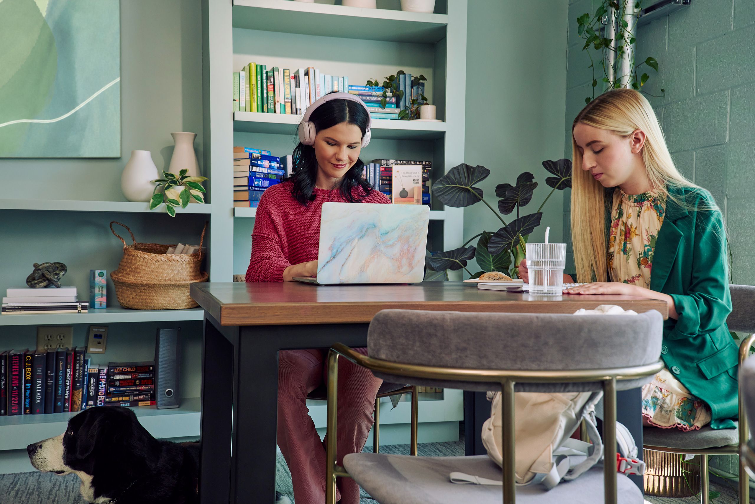 A young pale white blind woman and a young white woman with partially paralyzed arms and hands are coworking at a table in a modern coworking office space. The blind woman types on a computer while the woman with partially paralyzed arms and hands writes in a notebook. A glass of water with a straw in it is nearby. The woman with partially paralyzed arms and hands has long straight blonde hair and is wearing a floral dress and green blazer. The blind woman has shiny black hair styled in waves and is wearing a chunky knitted pink sweater and dusky pink pants. Her black and white guide dog is near her feet. The coworking office space is painted a warm mint green and features a large bookcase with several books, plants, clay vases, and a large abstract art piece.