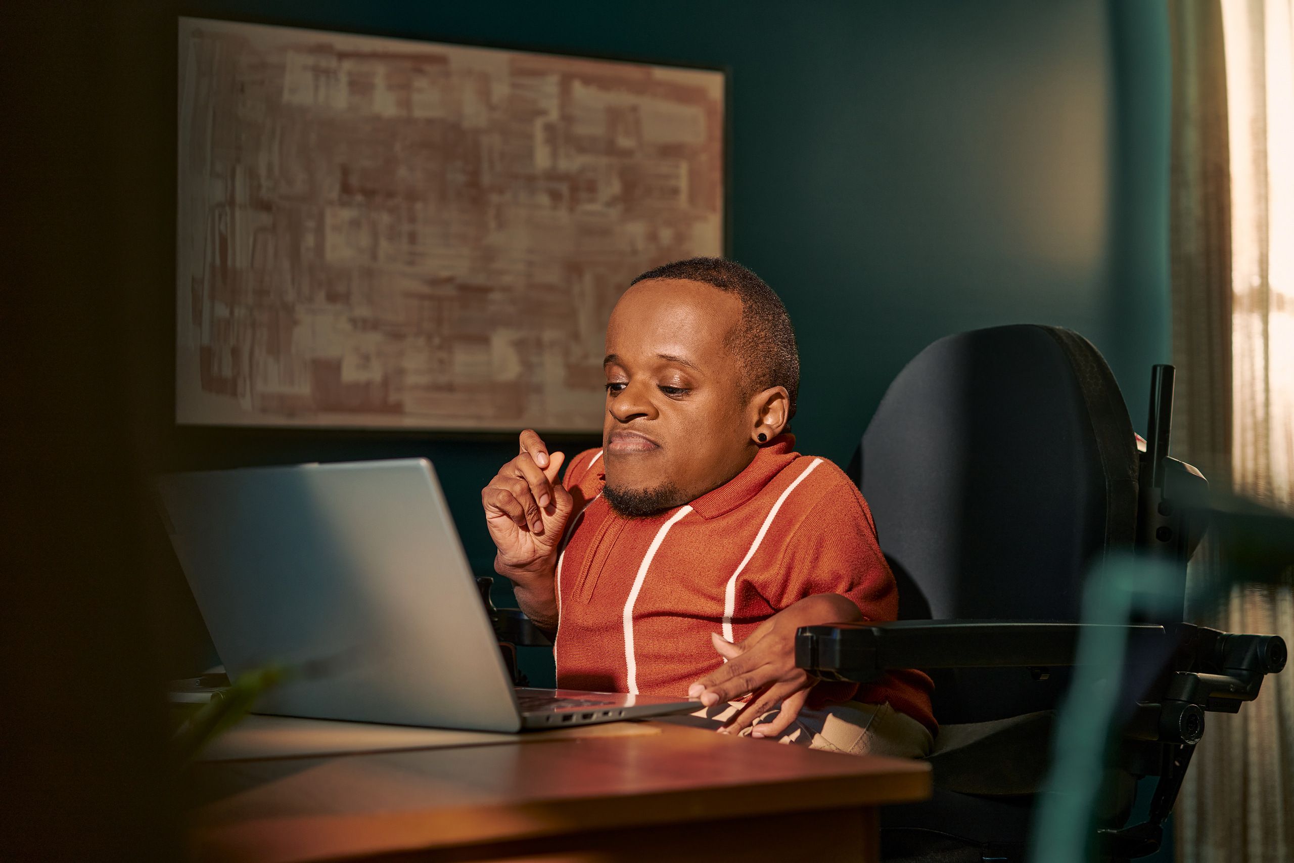 A black man who is a little person is seated in his power wheelchair in a dark teal home office. He types on a laptop on his desk, his hand held up in concentration.  An abstract art piece is on the wall behind him. He has closely cropped black hair, brown eyes, and is wearing a rust-orange knit quarter-zip sweater with white vertical stripes, light khaki trousers, and a small stud earring.