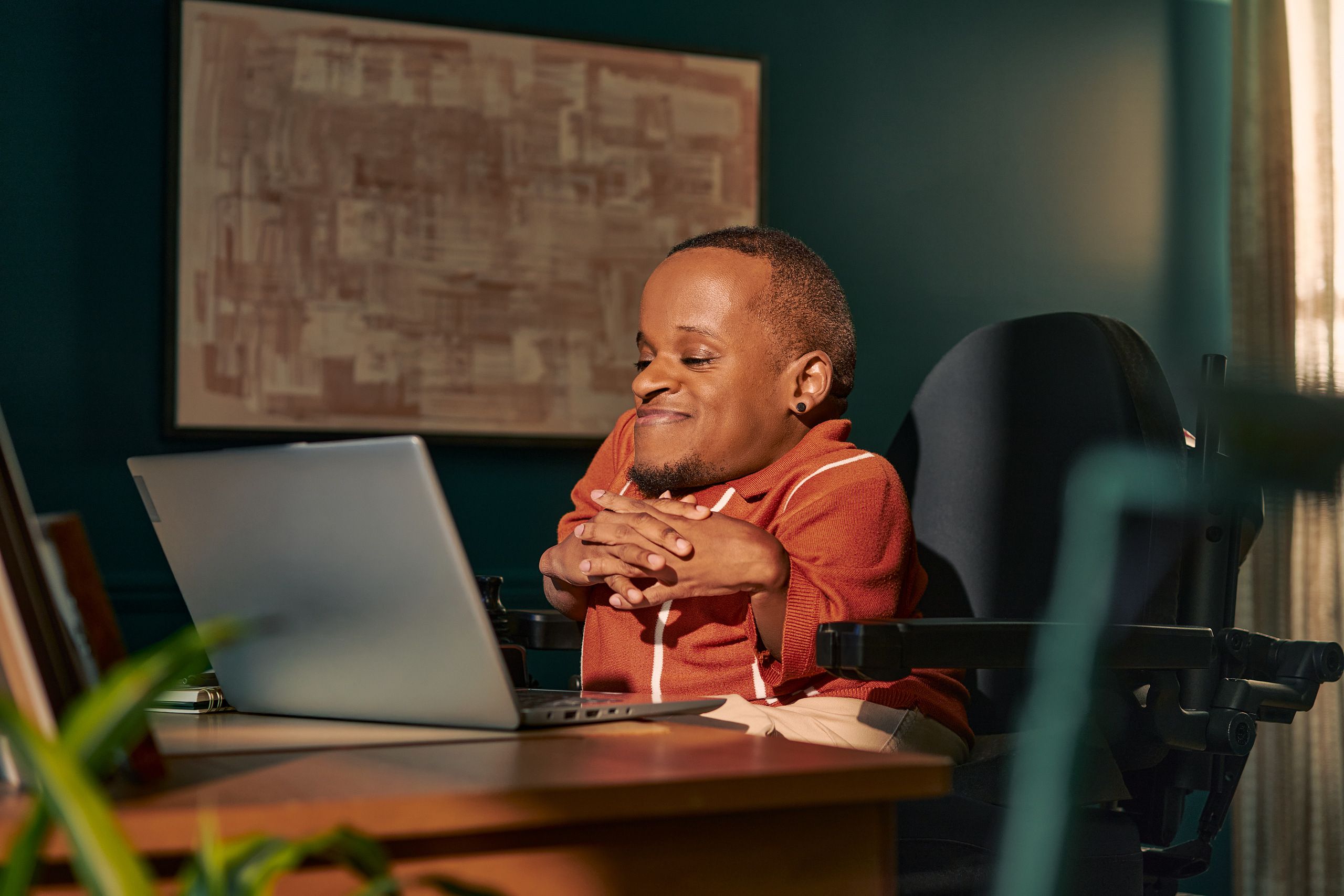 A black man who is a little person is seated in his power wheelchair in a dark teal home office. He smiles down at his open laptop, his hands together with fingers interlaced. A  notebook and framed photos are on the desk in front of him. An abstract art piece is on the wall behind him. He has closely cropped black hair, brown eyes, and is wearing a rust-orange knit quarter-zip sweater with white vertical stripes, light khaki trousers, and a small stud earring.