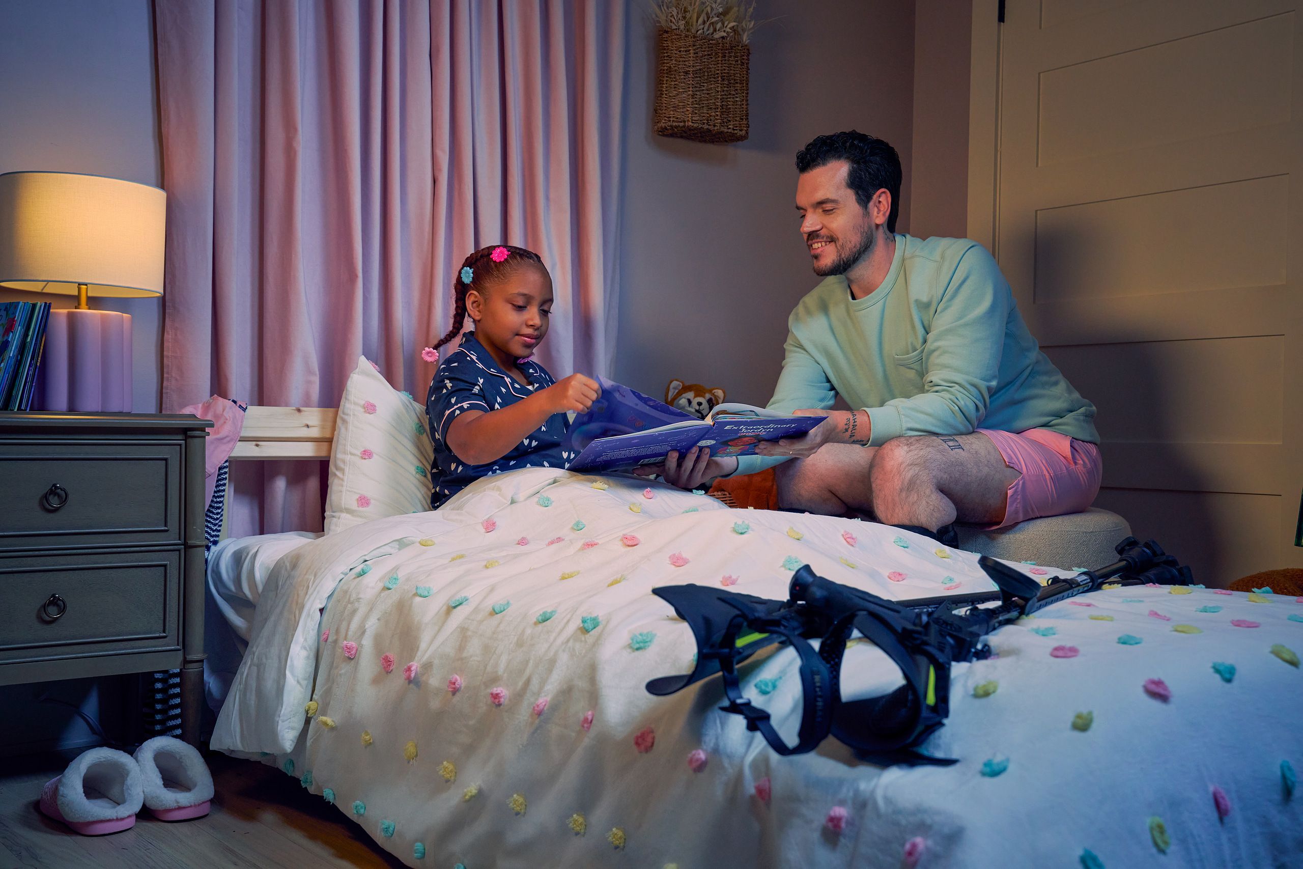 A white man with Cerebral Palsy who wears leg braces holds a children's book titled "Extraordinary Jordyn and Her Bionic Ears" to his daughter in her bedroom at night. His daughter is a Black multiracial American girl with thick braided red hair adorned with two flower barrettes and is wearing blue patterned pajamas. She is tucked into bed, holding a plushie animal and turns a page in the book. The father's black forearm crutches lie on the foot of the bed. He has dark brown curly hair, short brown facial hair, and is wearing a light green sweater with pink shorts. He has a tattoo of the Roman numeral two followed by a slash and the number three. The child's room is painted light pink and includes a nightstand, lamp, a pair of slippers, curtains, and children's books. Her bedspread is white with blue, pink, and yellow polka dots. Framed illustrations of Black women hang on the wall.