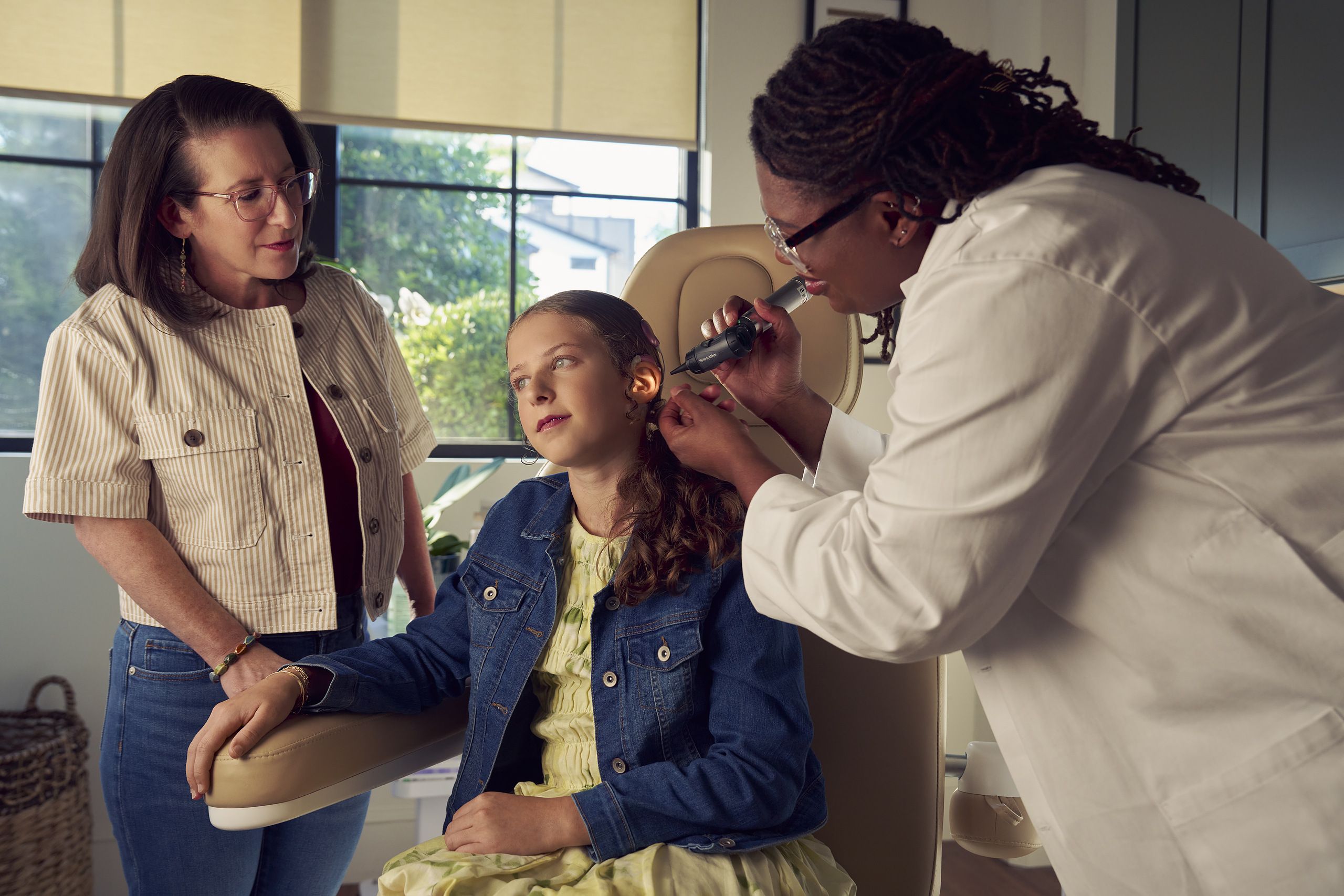 A Black deafBlind audiologist with a cochlear implant and glasses smiles and uses an otoscope to look into the ears of a young white deaf teen with braces seated in a medical treatment chair. The teen is softly smiling and has long, wavy sandy blonde hair and is wearing a jean jacket, and a yellow blouse. The teen's mother is standing next to her, observing the audiologist use the tool.
