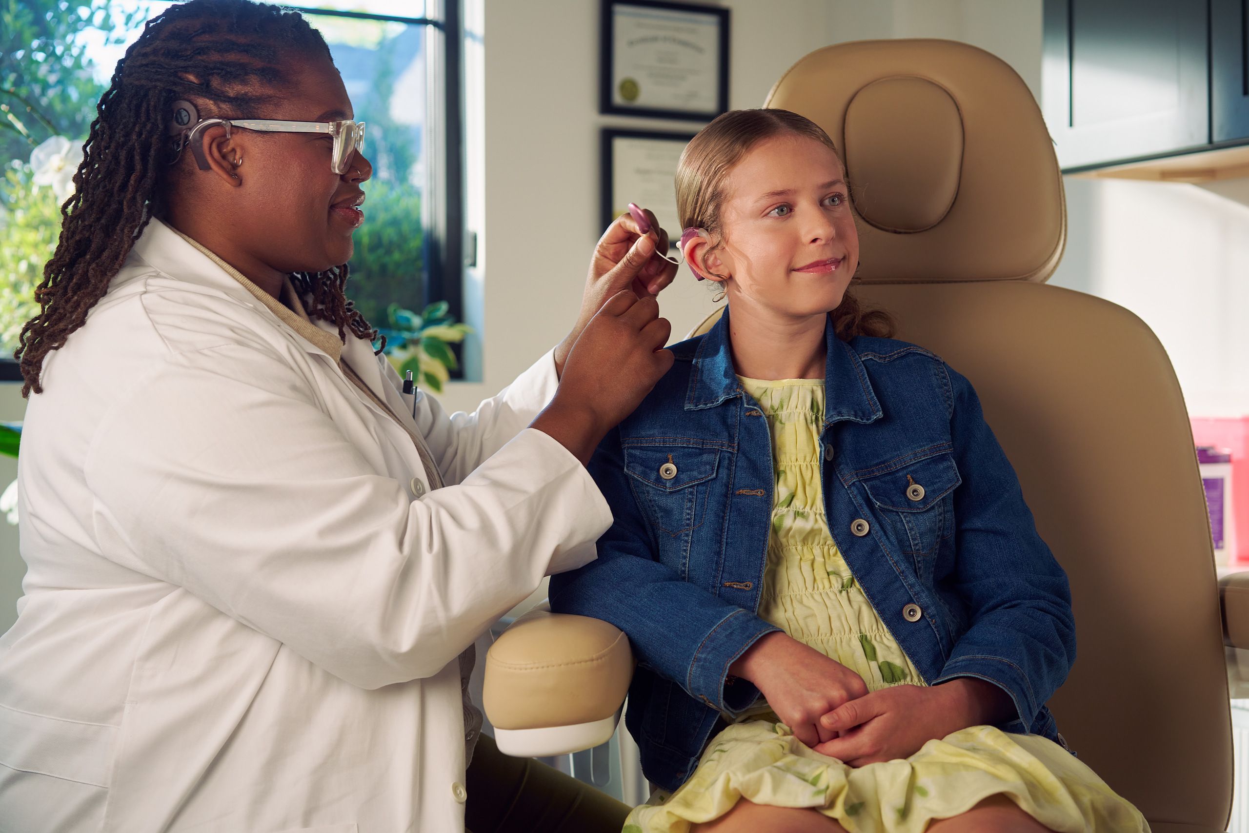 A Black deafBlind audiologist with a cochlear implant and glasses places a cochlear implant on a young white deaf teen with braces seated in a medical treatment chair. The teen is smiling and has long, wavy sandy blonde hair and is wearing a jean jacket, and a yellow blouse and skirt. Framed professional degrees are hanging on the wall behind them.