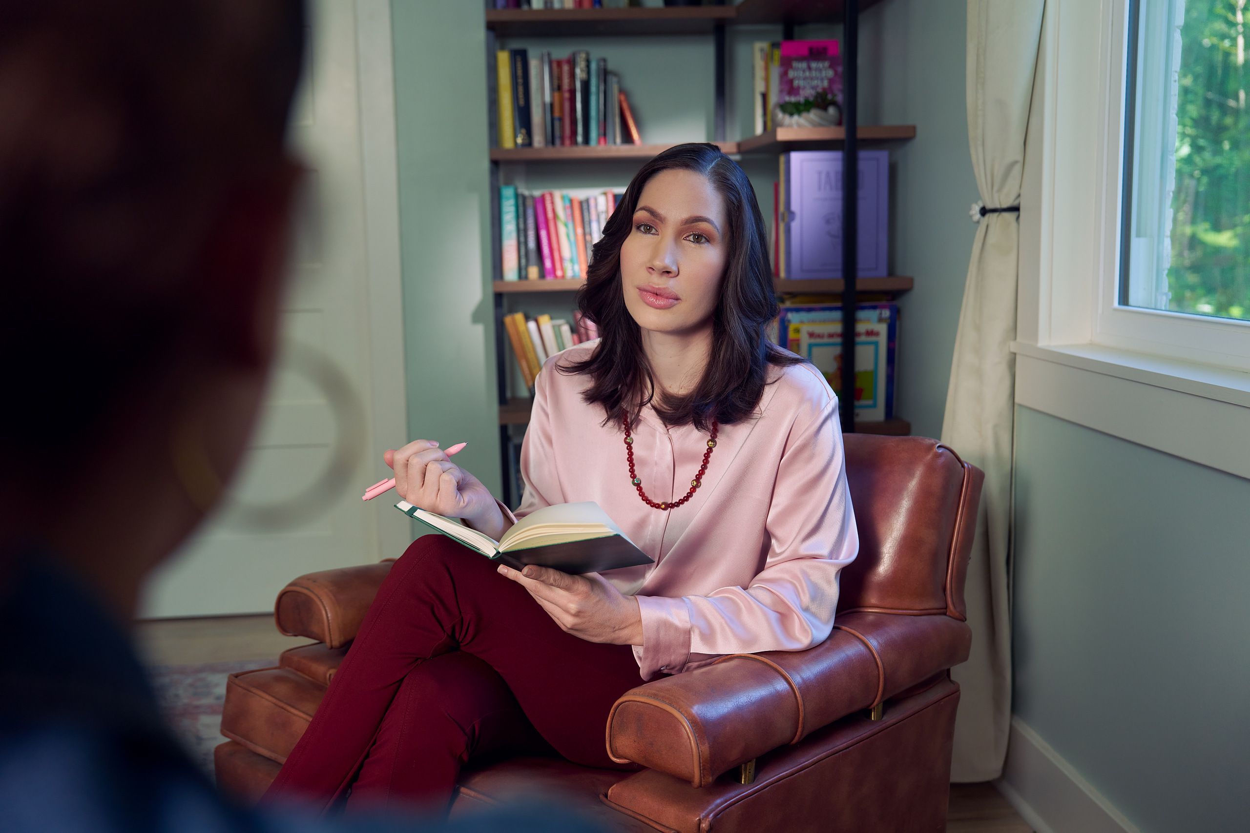 An Eastern European white woman therapist with a facial scar  sits in a large chair with an understanding look on her face. She is holding a pen and open notebook, and is looking at a client off camera. The therapist's office has several books on display and a window letting in natural light. The therapist is wearing a silk pink top, maroon dress pants, and a minimalist necklace.