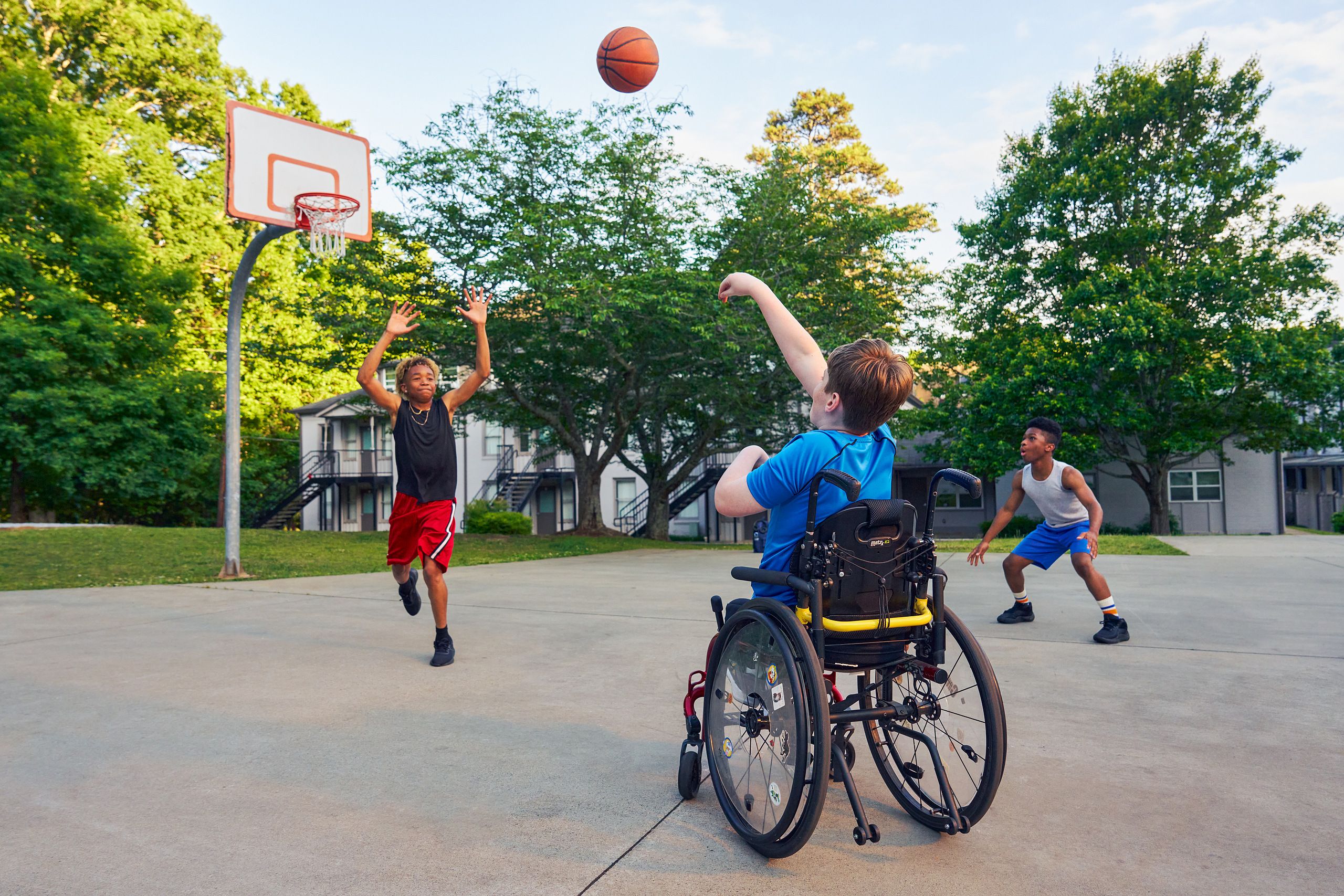 Two teens play basketball at a neighborhood park. One teen, a white boy seated in a manual wheelchair, shoots the ball. The other teen, a light-skinned African American male, jumps forward with arms outstretched, defending the shot. The teen seated in a manual wheelchair has short brown hair and is wearing blue athletic wear. The other teen is wearing black and red athletic wear.