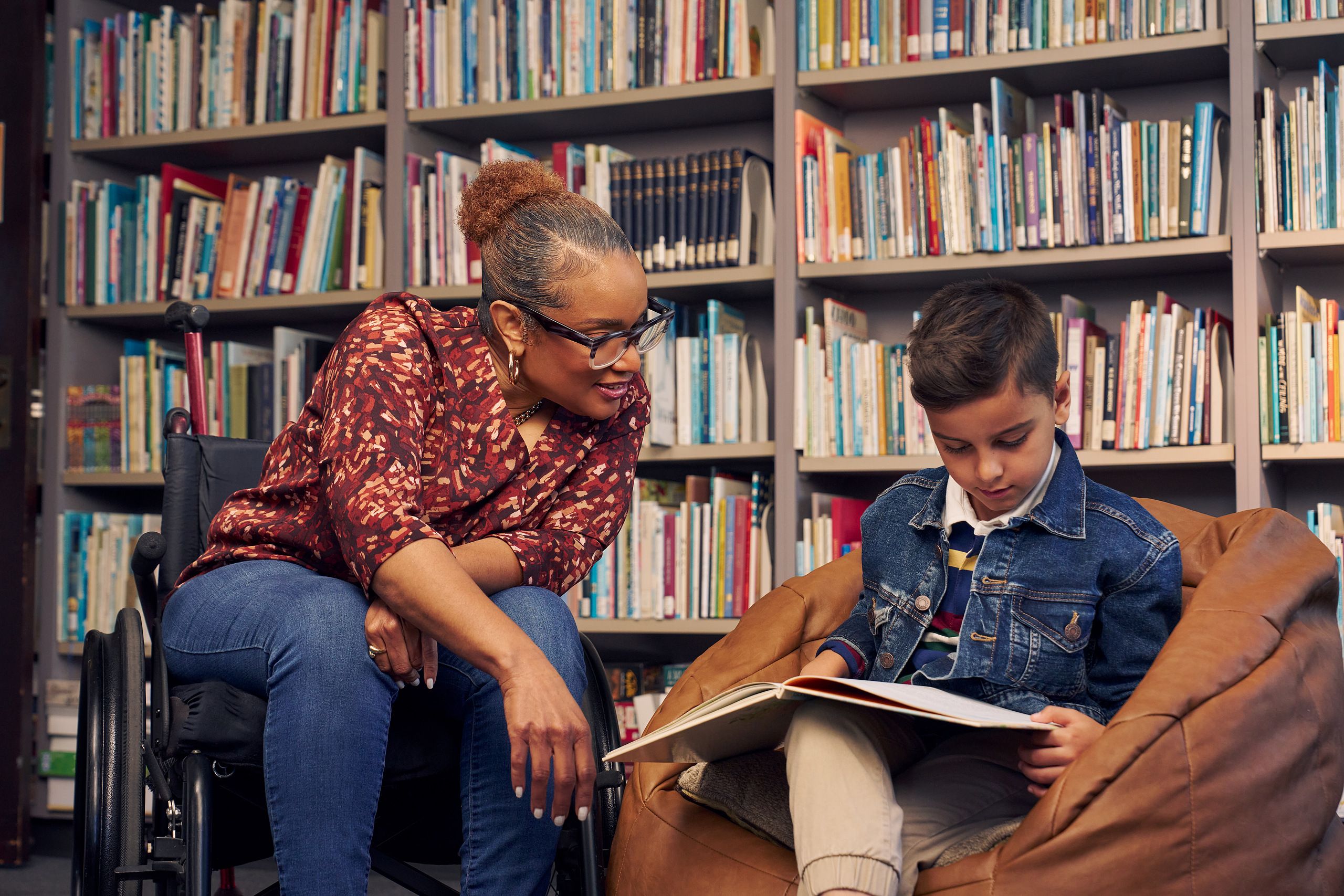 An African American teacher seated in a manual wheelchair smiles and watches a young student sitting in a bean bag chair read a picture book. The teacher has medium brown skin, reddish brown hair pulled into a short natural ponytail, and is wearing reading glasses, a red patterned blouse, and jeans. The young student is a Hispanic and Mediterranean-American boy with short brown hair. He  has olive skin and is wearing a blue striped shirt, a jean jacket, and khaki pants. A wall of children's books are behind them.