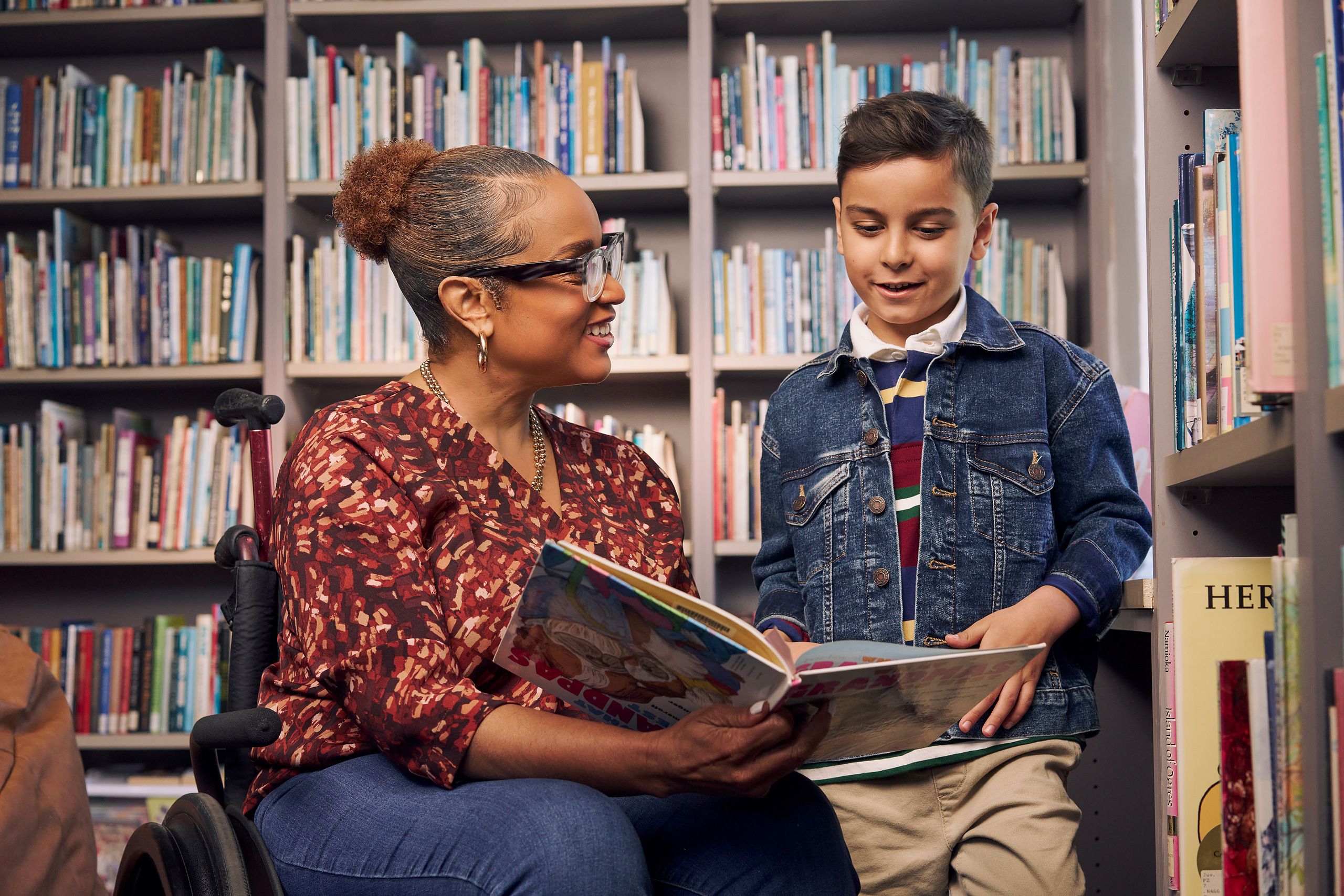 An African American teacher seated in a manual wheelchair smiles and holds a picture book open while her young student reads out loud. The teacher has medium brown skin, reddish brown hair pulled into a short natural ponytail, and is wearing reading glasses, a red patterned blouse, and jeans. The young student is a Hispanic and Mediterranean-American boy with short brown hair. He  has olive skin and is wearing a blue striped shirt, a jean jacket, and khaki pants. A wall of children's books are behind them.