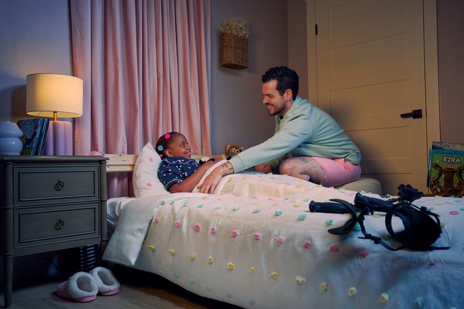 A white man with Cerebral Palsy who wears leg braces tucks his daughter into bed. His daughter is a Black multiracial American girl with thick braided red hair adorned with two flower barrettes and is wearing blue patterned pajamas. She is smiling in bed, and has several plushie animals nearby. The father's black forearm crutches lie on the foot of the bed. He has dark brown curly hair, short brown facial hair, and is wearing a light green sweater with pink shorts. He has a tattoo of the Roman numeral two followed by a slash and the number three. The child's room is painted light pink and includes a nightstand, lamp, a pair of slippers, curtains, and a stack of children's books. Her bedspread is white with blue, pink, and yellow polka dots.